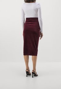 Burgundyröd sammets-midi kjol med en figurnära silhuett och draperingar, kombinerad med en vit långärmad topp och svarta högklackade sandaler.