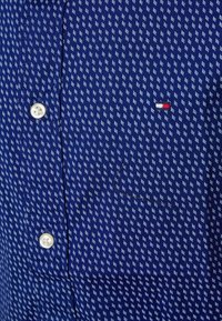 Marineblaues Hemd mit weißen und hellblauen geometrischen Mustern, ausgestattet mit weißen Knöpfen und einem kleinen Detail in Form eines Logos in Rot, Weiß und Blau.