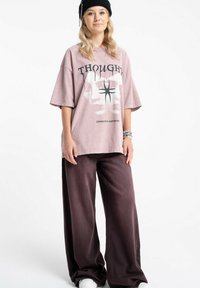 Młoda osoba w czarnej czapce typu beanie, dużym jasnofioletowym T-shircie z grafiką, szerokich ciemnofioletowych spodniach i białych trampkach, stojąca ze swobodnie opuszczonymi rękami.