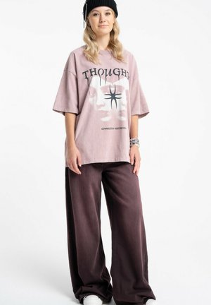 Młoda osoba w czarnej czapce typu beanie, dużym jasnofioletowym T-shircie z grafiką, szerokich ciemnofioletowych spodniach i białych trampkach, stojąca ze swobodnie opuszczonymi rękami.