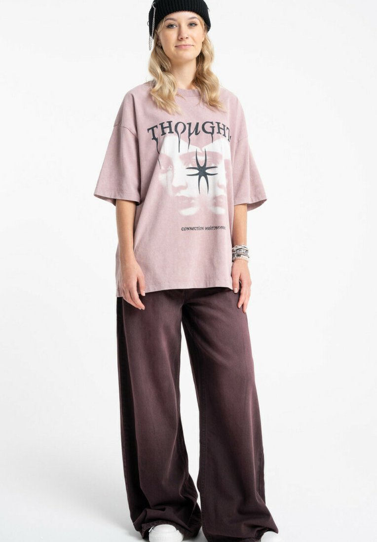 Młoda osoba w czarnej czapce typu beanie, dużym jasnofioletowym T-shircie z grafiką, szerokich ciemnofioletowych spodniach i białych trampkach, stojąca ze swobodnie opuszczonymi rękami.