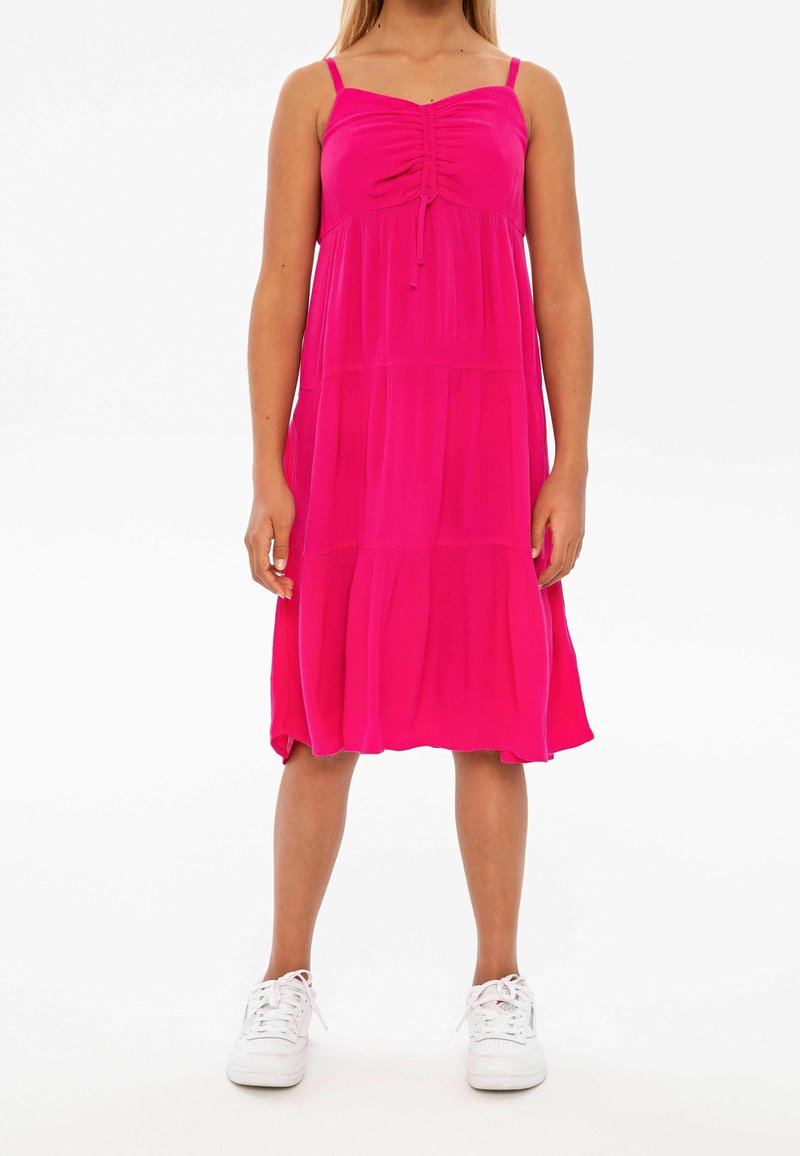 Femme portant une robe sans manches rose vif avec un devant froncé et des baskets blanches, debout devant un fond blanc uni.