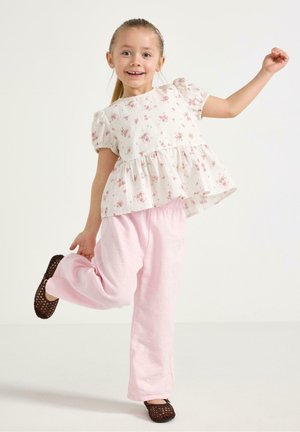 Ung pige smiler, balancerer på ét ben, iført en hvid bluse med blomsterprint, lyserøde bukser og brune perforerede sko.