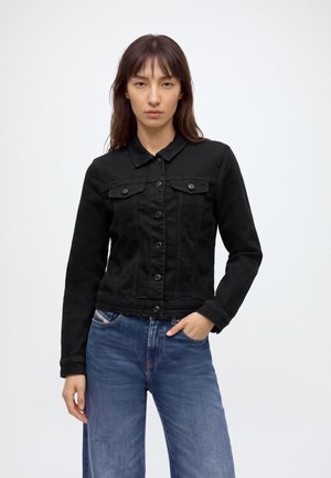 Femme aux longs cheveux foncés porte une veste en jean noire boutonnée et un jean bleu, debout avec une main dans la poche devant un fond uni.