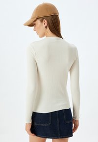 Wit, langmouwig ribbed top gecombineerd met een donkere spijkerrok. De outfit wordt geaccentueerd door een tan kleurige baseballcap.