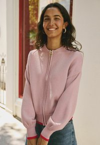 Světle růžový pletený kardigan s pružnými manžetami a spodním lemem, se zlatým zipem ve tvaru hvězdy. Kombinován s modrou džínsovou sukní.