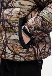 Polstret jakke med et skov camouflage mønster i brune, grønne og sorte nuancer. Inkluderer en lynlåslomme og justerbare manchetter.