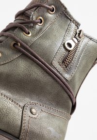 Zielony skórzany botek za kostkę z brązowymi sznurówkami, zamkiem błyskawicznym po boku i metalowymi oczkami; posiada przeszywane akcenty oraz detal z logo w pobliżu podeszwy.