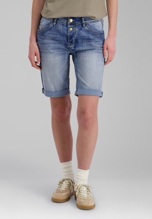 Person, die hochgekrempelte blaue Jeansshorts, beige Sneaker, weiße Socken und ein beigefarbenes Oberteil trägt, steht vor einem schlichten grauen Hintergrund.