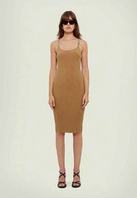 Mulher vestindo um vestido midi sem mangas, canela e com alças com fivela, saltos pretos com tiras e óculos de sol escuros, em pé contra um fundo simples.