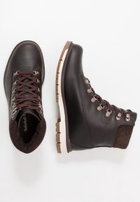 Botas de senderismo de cuero marrón con un collar de ante, cordones redondos, ojales de metal y una media suela blanca. La suela exterior presenta patrones de agarre texturizados.