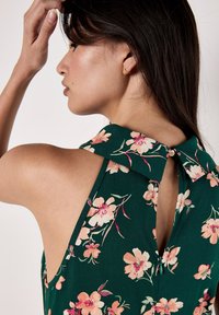 Haut halter fleuri en tissu vert foncé avec des fleurs roses et crème. Les détails remarquables incluent un col et un dos en trou de serrure avec fermeture par bouton.