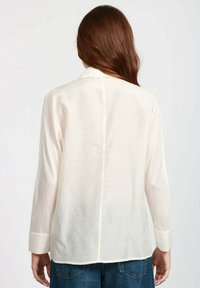 Camicia a maniche lunghe color beige chiaro in un tessuto morbido e trasparente, con taglio dritto, colletto e polsini con bottoni, caratterizzata da un dettaglio di cucitura sul retro.