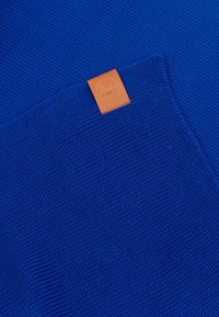 Niebieska dzianina o teksturowanej powierzchni, z kwadratową brązową skórzaną metką w dolnym rogu z wytłoczonym logo.