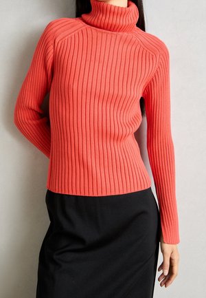 Kobieta w koralowym prążkowanym golfie z długimi rękawami i czarnej spódnicy, stojąca na tle jednolitego jasnego tła.