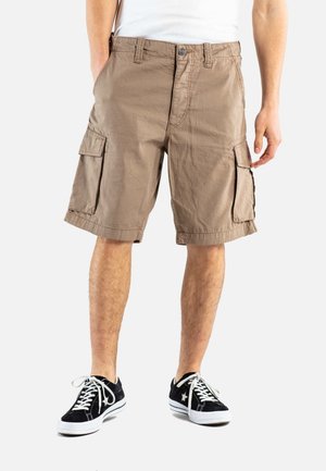 Man draagt beige cargoshorts en zwarte lage sneakers met witte veters, staand tegen een effen witte achtergrond.