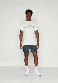 Hombre de pie contra un fondo neutro, vistiendo una camiseta blanca con el lema "Just Do It", pantalones cortos oscuros, calcetines blancos y zapatillas blancas.