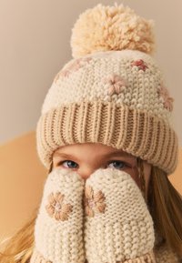 Strikket beige hat med pom-pom og blomsterbroderi på en cremefarvet baggrund. Matchende vanter dækker ansigtet og viser et lignende blomsterdesign.