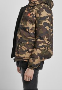 Kurtka pikowana w kamuflażu w odcieniach zieleni, brązu i beżu, z zamkiem błyskawicznym, elastycznymi mankietami oraz pomarańczową naszywką z logo. Z przodu znajdują się kieszenie.