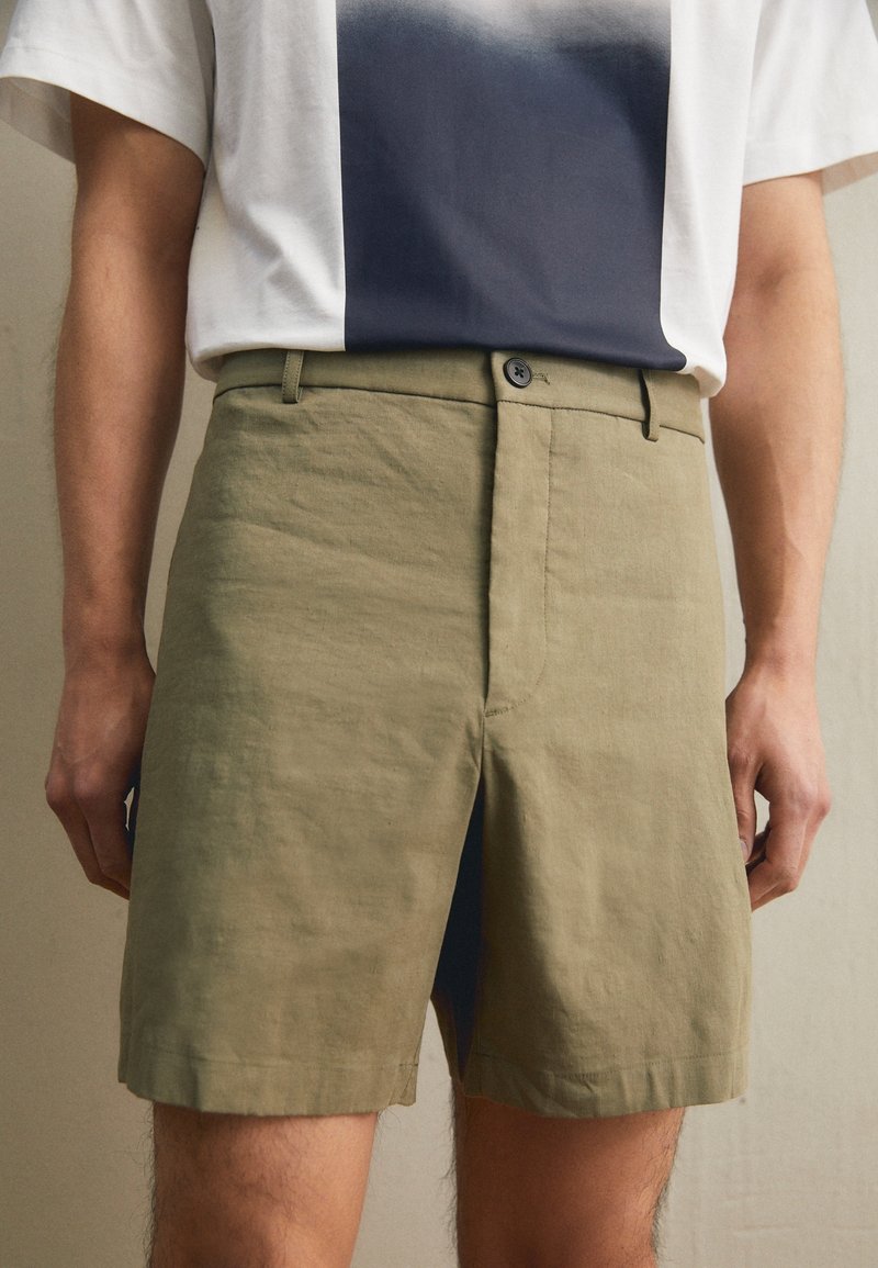 Homme portant un short beige jusqu'aux genoux et un t-shirt blanc avec un grand motif carré foncé à l'avant, debout avec les bras détendus.