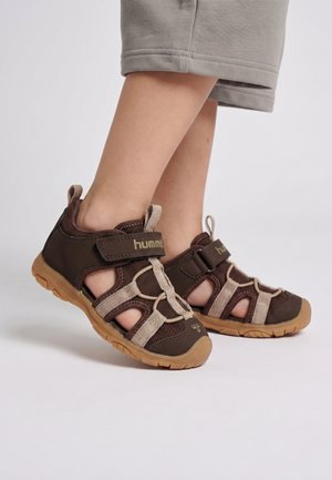 Child wearing brown and beige closed-toe sandals with velcro straps and rubber soles, standing on a white surface.