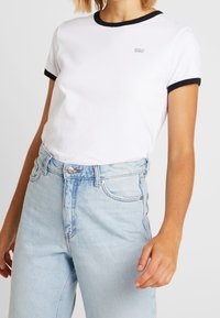 Vit bomull t-shirt med svart ringkrage och manschetter; har en liten logotyp på bröstet; matchar med ljusblå hög midjejeans.