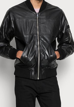 Person trägt eine schwarze Leder-Bomberjacke mit gerippten Bündchen und Saum, Frontreißverschluss, Seitentaschen und einer Reißverschlusstasche am Ärmel.