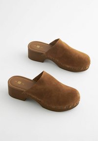 Bruine suède klompen met een ronde neus en een gladde hak. Beschikt over decoratieve metalen studs aan de zijkant. Vlakken houten zool.