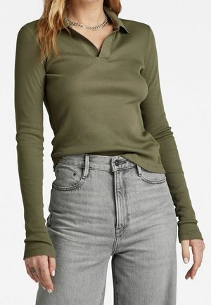 Kobieta w oliwkowej, prążkowanej koszulce polo z długim rękawem, wpuszczonej w jasnoszare jeansy z wysokim stanem, ukazująca górną część tułowia i dłonie.