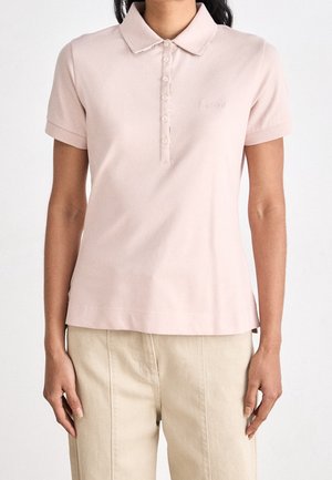 Kobieta w jasnoróżowej koszulce polo z krótkim rękawem i guzikami oraz beżowych spodniach, stoi z ramionami swobodnie opuszczonymi wzdłuż boków.