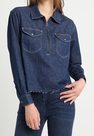 Giacca corta in denim scuro con chiusura frontale con zip, due tasche sul petto e orlo sfilacciato. Dotata di colletto classico e maniche lunghe.