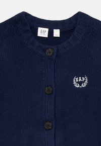 Modra pletena jopica z rebrasto teksturo, ki ima okroglo ovratnik, štiri črne gumbe in belo vezeno logo GAP z lovorjevim oblikovanjem.