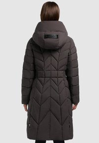 Mujer vestida con un abrigo de invierno acolchado largo y oscuro, con una gran capucha, vista desde atrás sobre un fondo liso.