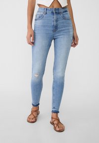Jeans skinny de mezclilla azul claro con detalles desgastados en las rodillas, cintura alta y un ajuste hasta el tobillo, combinados con sandalias marrones de tiras.