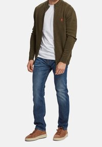 Uomo che indossa un cardigan verde oliva con zip sopra una maglietta bianca, jeans blu e sneakers in camoscio marroni, in piedi contro uno sfondo bianco.