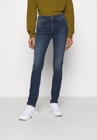 Osoba oblečená v tmavě modrých slim jeans, bílých teniskách a dlouhém olivově zeleném topu, stojící na pozadí bez vzoru bílé barvy.