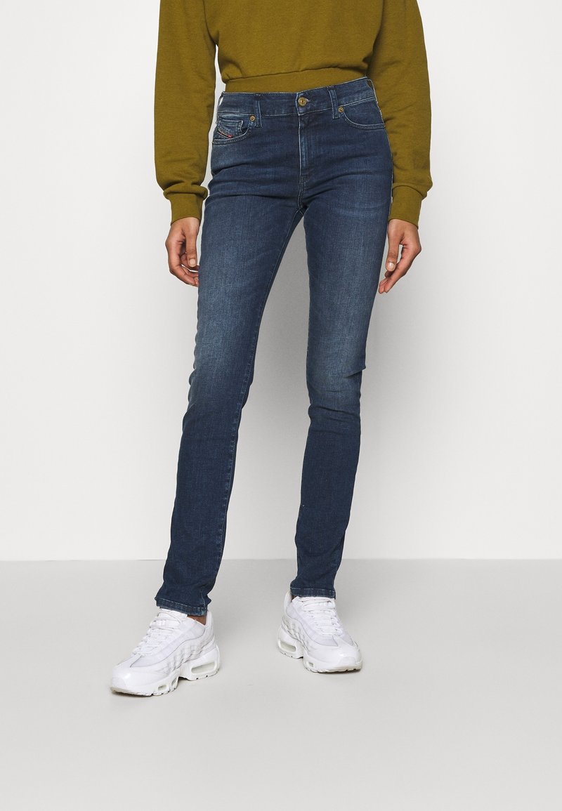 Osoba oblečená v tmavě modrých slim jeans, bílých teniskách a dlouhém olivově zeleném topu, stojící na pozadí bez vzoru bílé barvy.