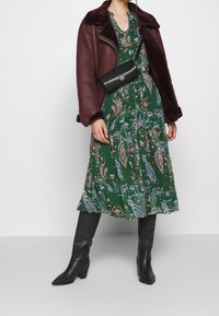 Mujer con un vestido verde de paisley, chaqueta de borreguillo burdeos, bolso cruzado negro y botas altas de tacón negro.