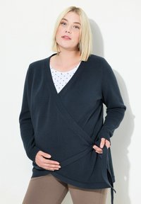 Tmavě modrý zavinovací svetr s výstřihem do V, strukturovaným ripem a bočním zavazováním, nošený přes bílé tričko s puntíky a kombinovaný s hnědými legínami.