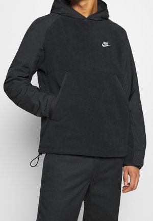 Person iført sort Nike hættetrøje med forlommer og hætte, sorte bukser, stående mod en ensfarvet baggrund.