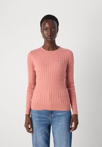 Pulover tricotat roz cu un design cu cabluri, decolteu rotund și mâneci lungi. Asortat cu blugi din denim bleu deschis. Detaliu simplu cu logo pe piept.