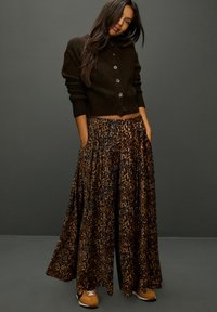 Cropped black cardigan with buttons, paired with a flowing brown leopard print maxi skirt featuring pockets. Brown sneakers complete the outfit.