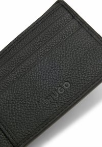 Porte-carte en cuir noir avec une finition texturée, quatre emplacements pour cartes et un logo embossé. Le design est épuré et minimaliste.