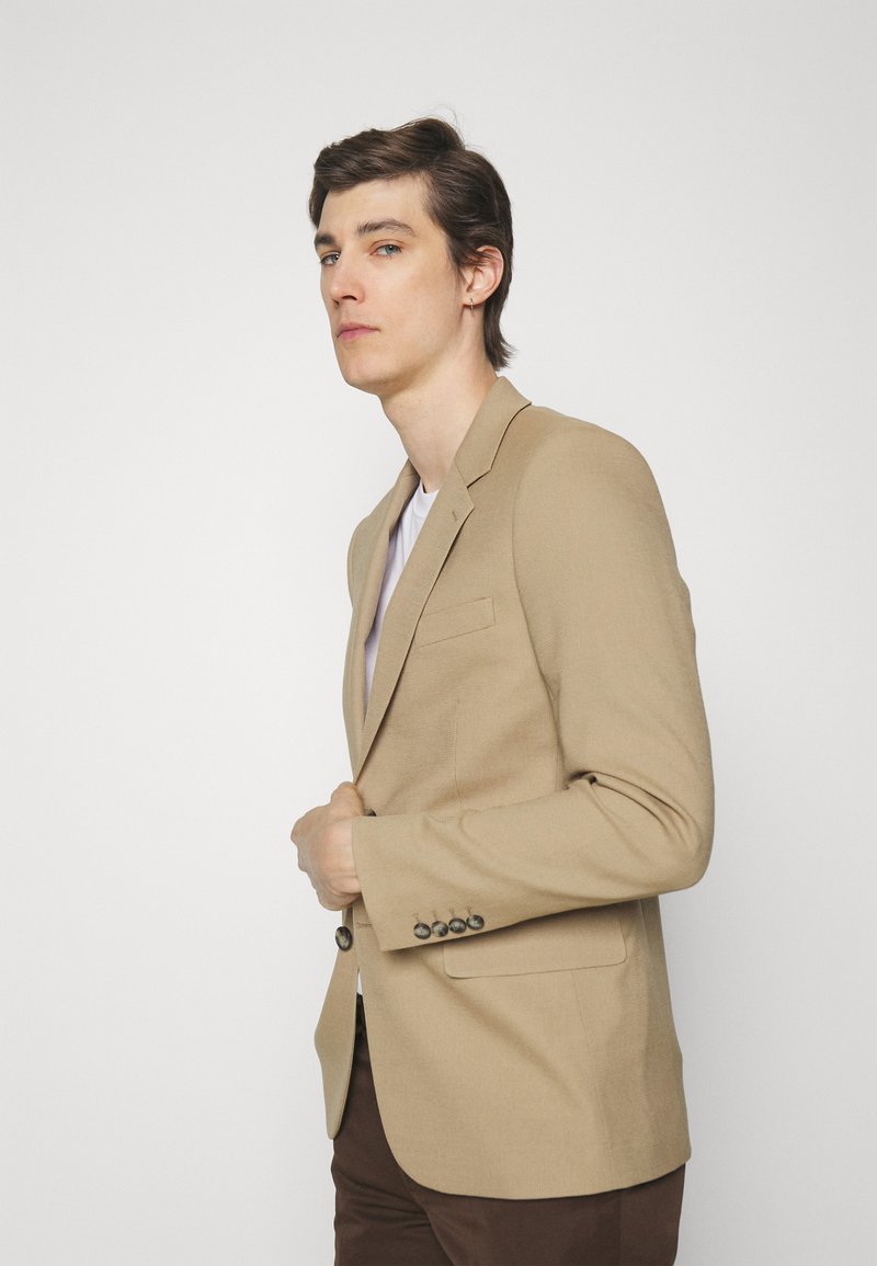 Light brown blazer made of textured fabric, featuring a slim fit, single-button closure, and a pocket on the left side.