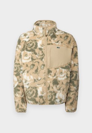 Fuzzy fleece jacket in beige with green floral pattern, featuring a zip front and a tan chest pocket. Lacoste logo on pocket.
