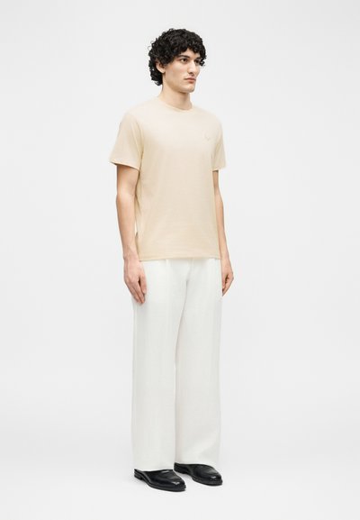 Άνδρας με σγουρά σκούρα μαλλιά που φοράει μπεζ κοντομάνικη μπλούζα, λευκό παντελόνι με φαρδιές παντελόνες και μαύρα παπούτσια, στέκεται μπροστά σε απλό λευκό φόντο.