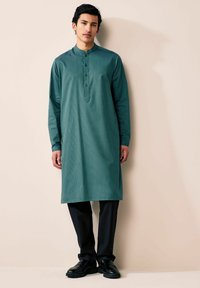 Turkusowa długa kurta z kołnierzykiem mandaryńskim, z guzikowym zapięciem. Połączona z czarnymi spodniami i czarnymi eleganckimi butami. Gładka tekstura.