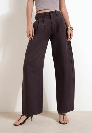 Person iført sort højtaljede bukser med vide ben, grå cropped top, gyldne armbånd og sorte hælede sandaler, stående på lys gulv.
