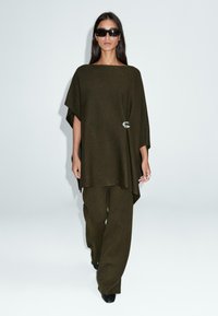 Donna che indossa pantaloni larghi verde oliva scuro e top abbinato largo con spilla argento, scarpe nere e grandi occhiali da sole neri, che cammina in avanti.