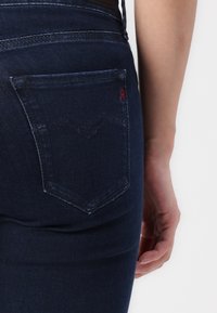 Jeans de mezclilla de color azul oscuro con un diseño ajustado, bolsillo trasero con un sutil logo rojo y ligeros detalles de desgaste en la tela.